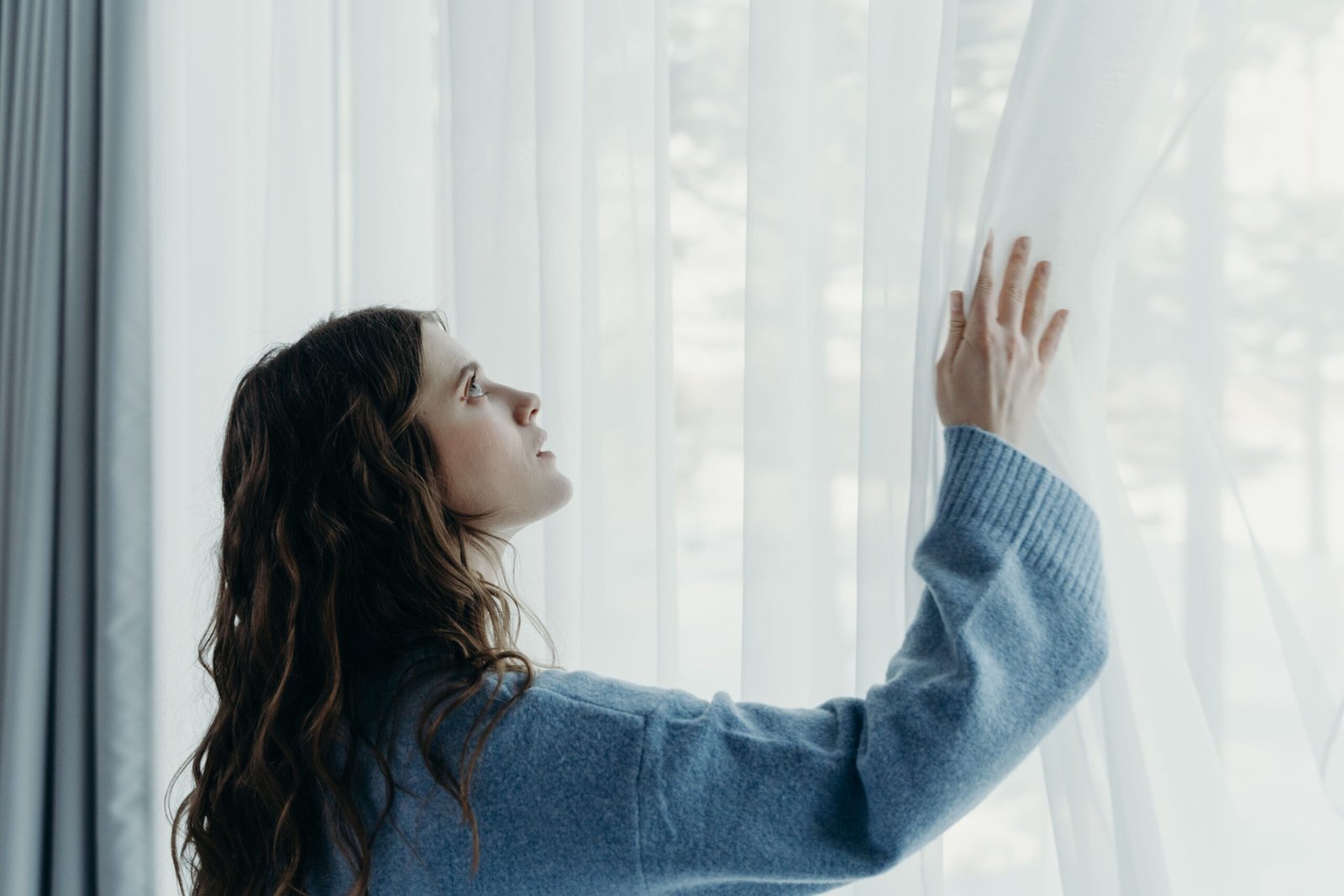 Woman in a blue sweater gently touches sheer curtains with natural light streaming in.